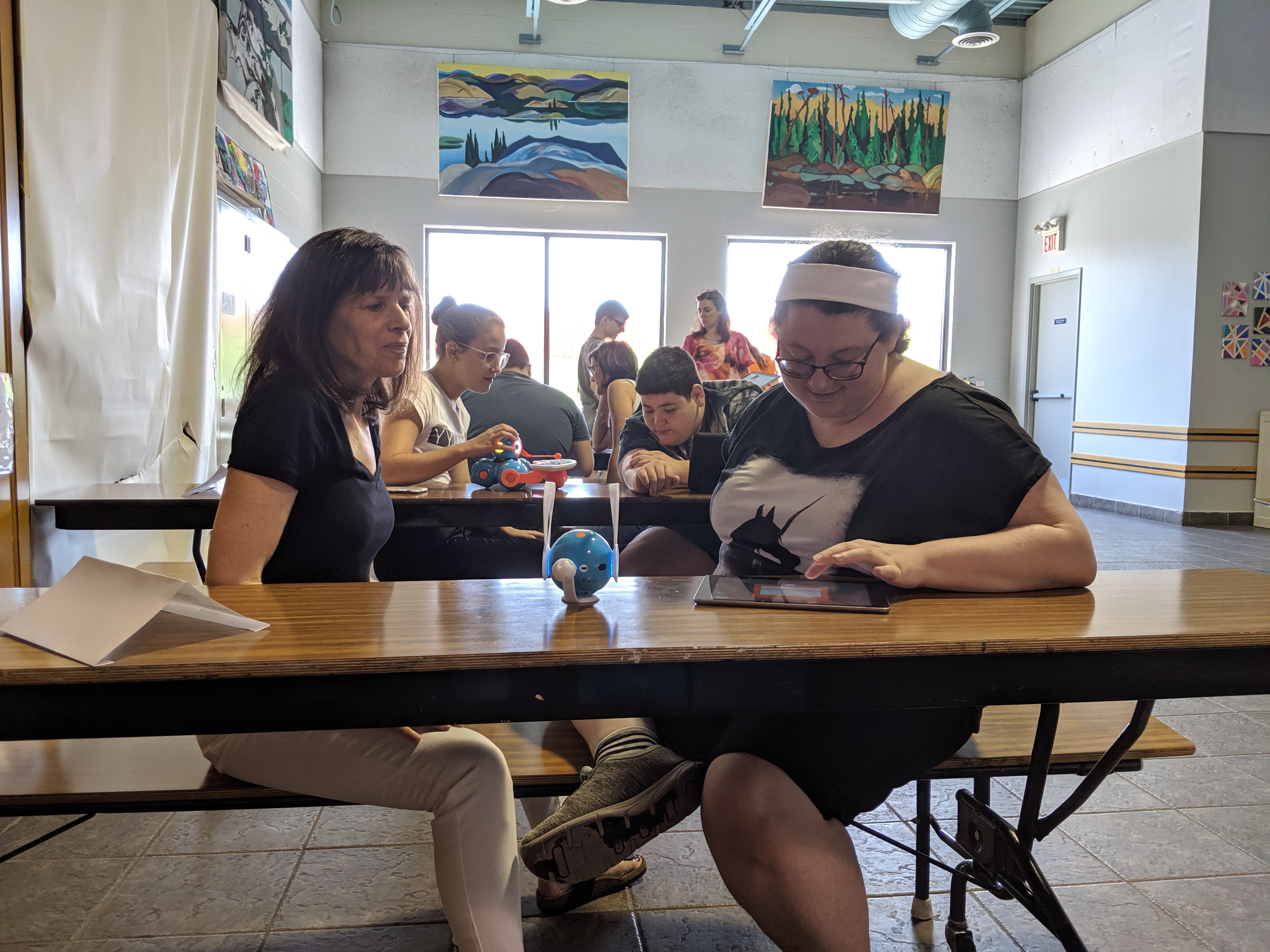 In a classroom environment, each facilitator is paired with a student sitting at a table. Each student is using an iPad to program a robot placed on the same table.